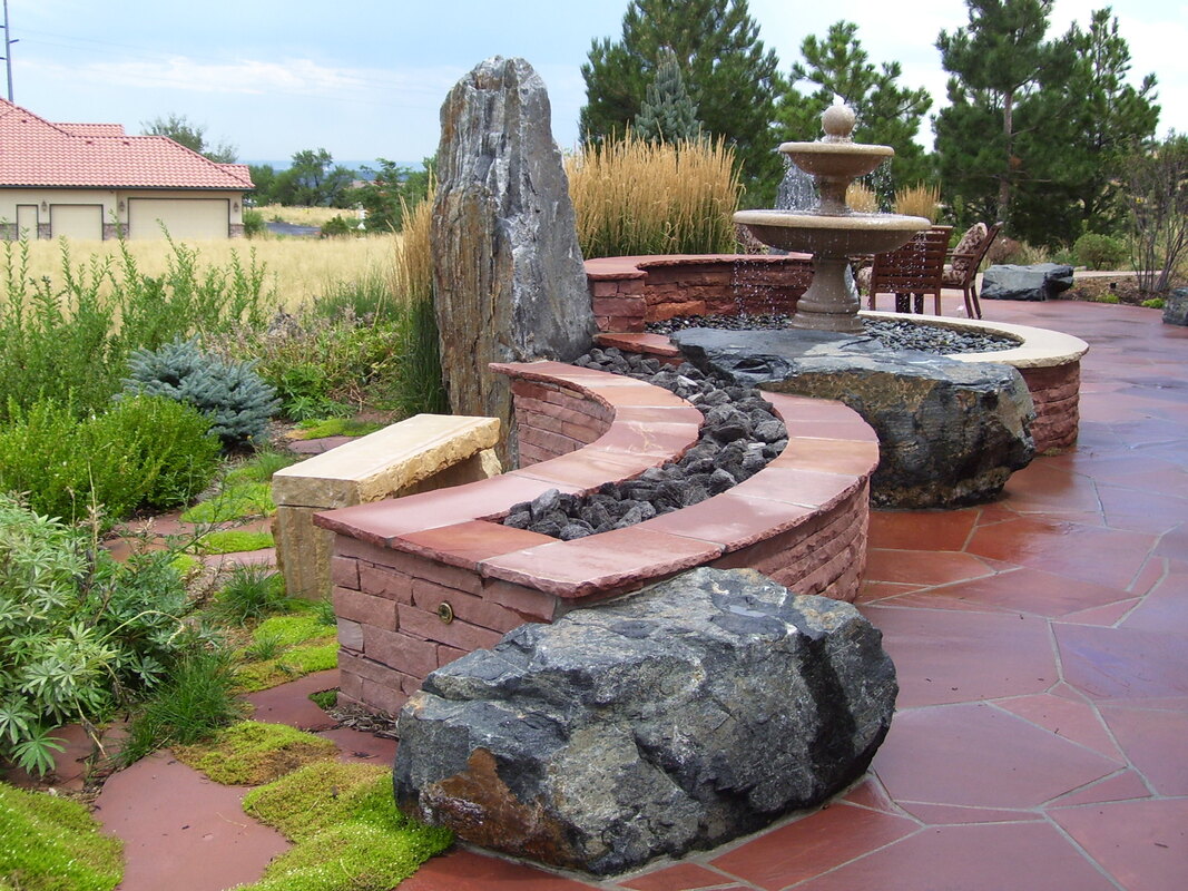 Picture of a Denver metro residential landscape design featuring a red stone patio with raised brick fire features and a self contained fountain. 